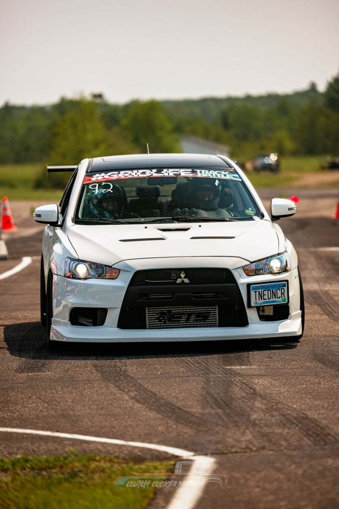 A white sports car drifts on a racetrack, leaving tire marks on the asphalt. Glitches and colorful visual artifacts add energy, as if the scene were a highlight from the Lake Superior Ice Festival. Blurred trees and scenery complete the background.
