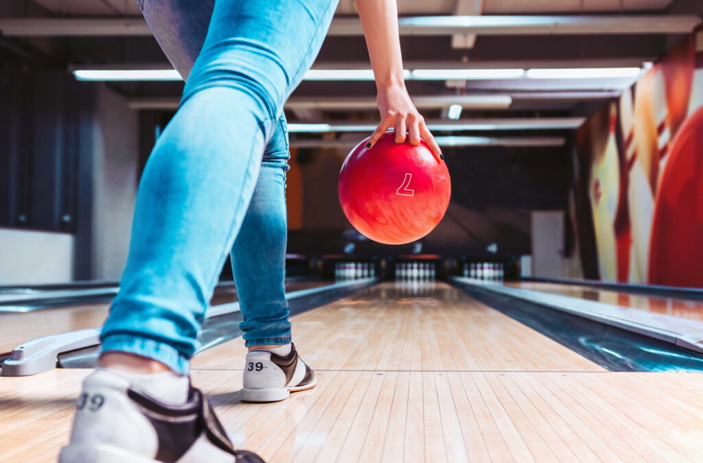 A person wearing jeans and bowling shoes is about to roll a red bowling ball down a lane at the Lake Superior Ice Festival. The image is taken from behind, focusing on the player's stance and the ball.