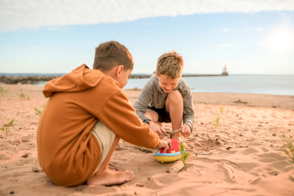 Two young boys sit on a sandy beach, crouched closely together, playing with a small toy boat—a scene that captures the charm of Wisconsin places to visit in summer, with the ocean and distant lighthouse under a blue sky.