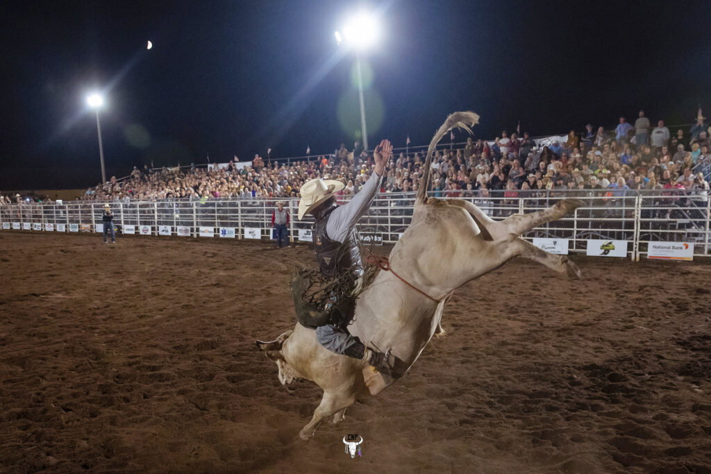 A rodeo rider wearing a hat and vest rides a bucking bull at night in a dirt arena, as a large crowd watches from bleachers under bright lights and a crescent moon, capturing the excitement of events like the Lake Superior Ice Festival.