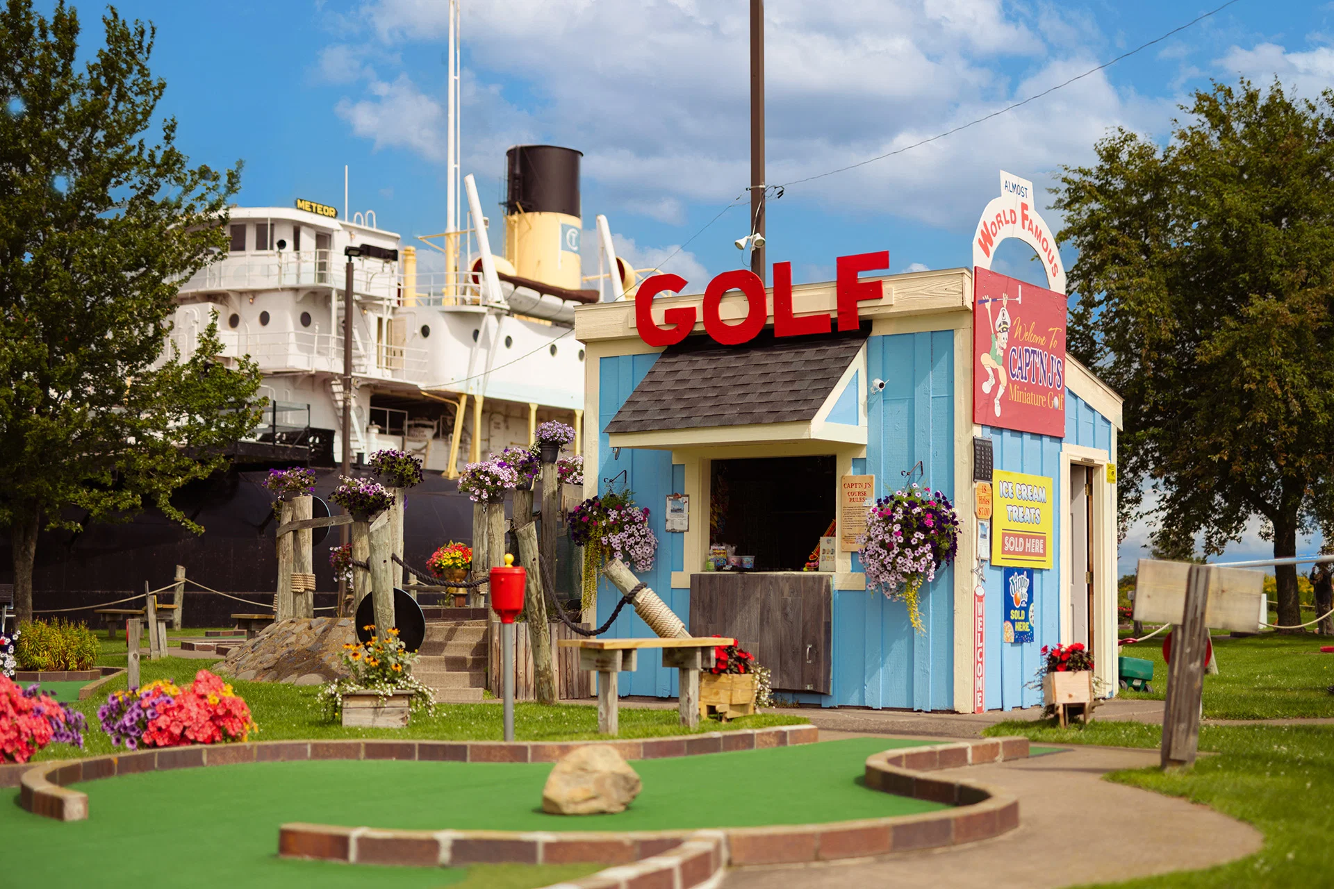 A colorful mini-golf kiosk with "GOLF" in red letters stands on a green course, surrounded by flowers—one of the fun Wisconsin places to visit in summer. A large ship is visible in the background under a partly cloudy blue sky.