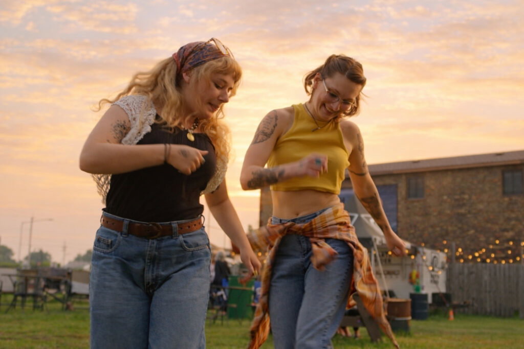 Two women laugh and dance together outdoors at sunset, enjoying a summer evening in jeans and casual clothes. With string lights and a building behind them, it's the perfect scene for discovering Wisconsin places to visit in summer.