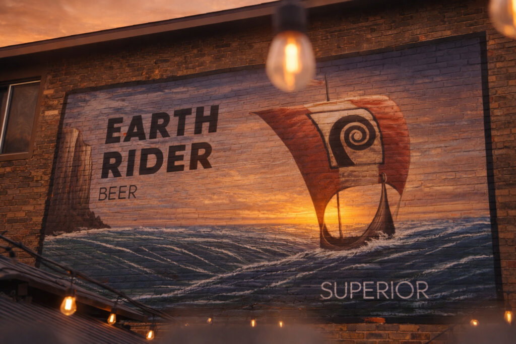 A mural on a brick wall shows a Viking ship sailing on waves at sunset, with the words "EARTH RIDER BEER SUPERIOR" painted beside it. Warm lights hang in the foreground, celebrating the spirit of the Lake Superior Ice Festival.