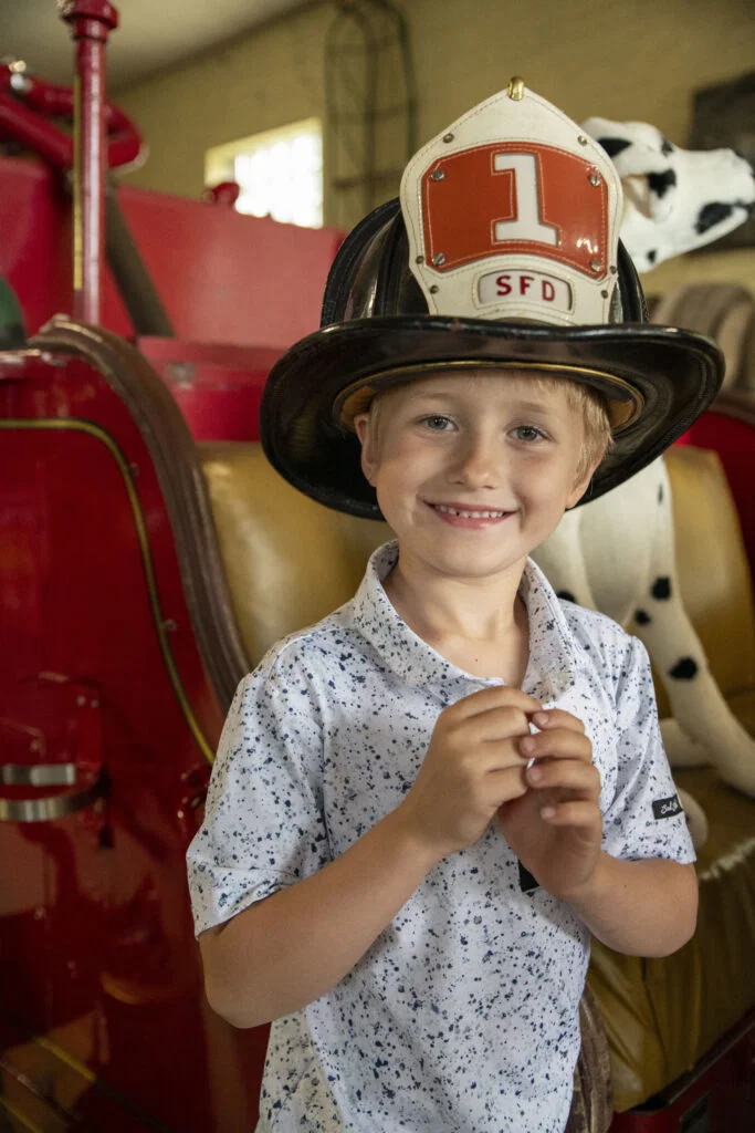 A smiling young boy wearing a large firefighter helmet with a red "1 SFD" badge stands in front of a vintage red fire truck and a plush Dalmatian—perfect for families exploring Wisconsin places to visit in summer.