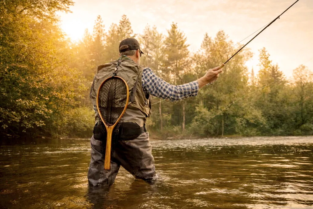 A person wearing waders and a vest stands in a river, fly fishing at sunset. Surrounded by trees and greenery, they enjoy one of the many outdoor activities in Superior Wisconsin, with a wooden fishing net hanging from their back.