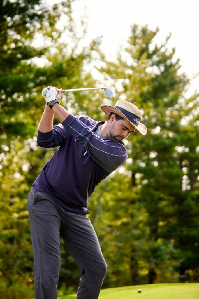 A man wearing a straw hat and a dark long-sleeve shirt prepares to swing a golf club on a green course, reminiscent of the festive spirit found at the Lake Superior Ice Festival, with trees and a bright sky in the background.