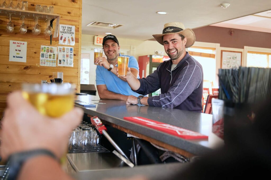 Two men sit at a bar during the Lake Superior Ice Festival, smiling and raising glasses of beer in a toast. One wears a cowboy hat and the other a baseball cap. Another person in the foreground also holds up a drink. The vibe is casual and friendly.