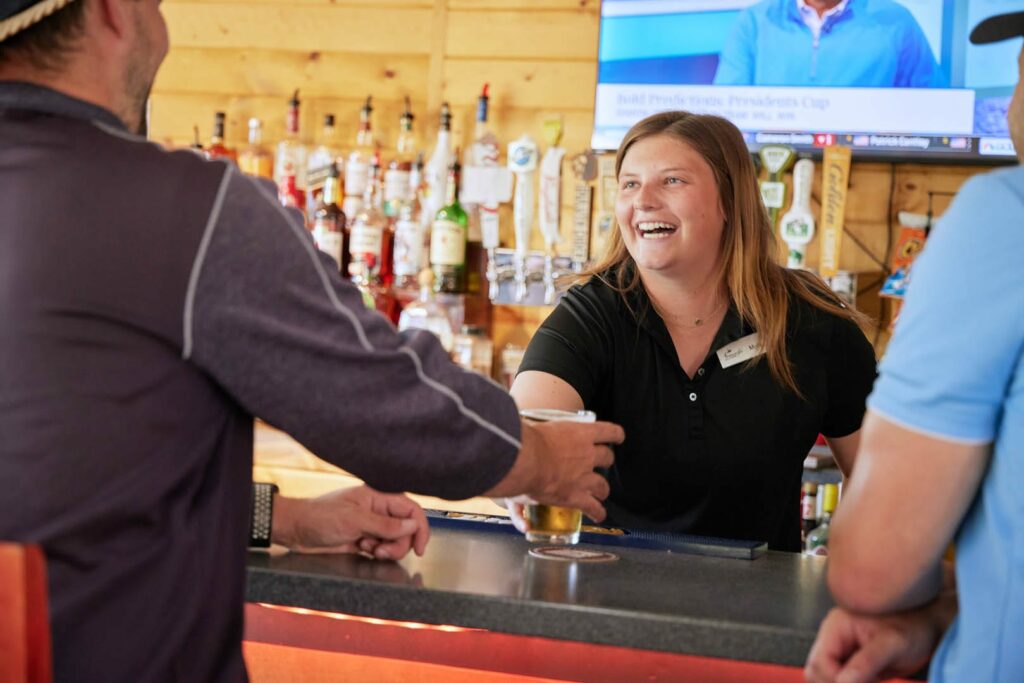 A smiling bartender hands a glass of beer to a customer at a bar during the Lake Superior Ice Festival, with bottles and taps behind her, two people sitting at the counter, and a TV on in the background.