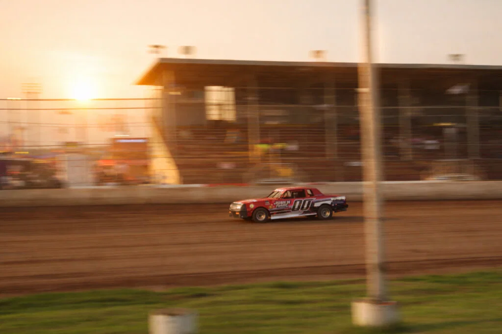 A red and white race car speeds along a dirt track at sunset, with blurred motion and an empty grandstand in the background—a thrilling scene reminiscent of classic Wisconsin places to visit in summer.
