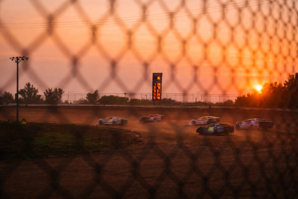 Race cars speed around a dirt track at sunset, viewed through a chain-link fence. A digital scoreboard displays lap counts in the background, with trees silhouetted against the orange sky.