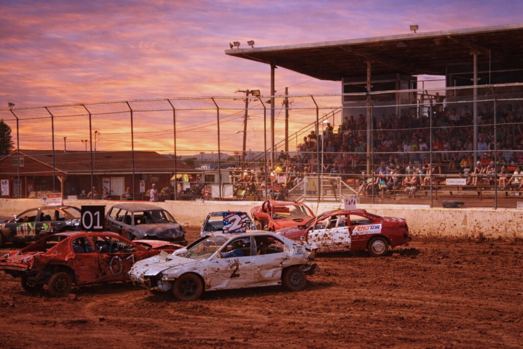 A group of battered cars compete in a demolition derby on a dirt arena at sunset during the Lake Superior Ice Festival, with a grandstand full of spectators watching behind a wire fence.