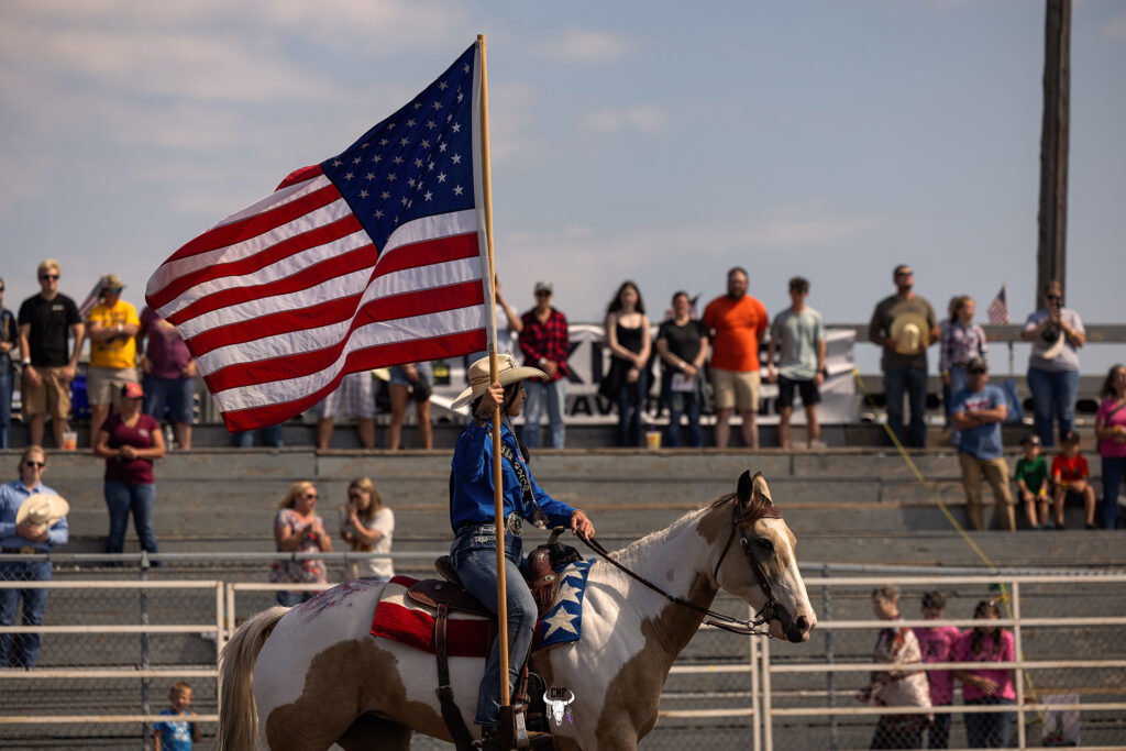 A person in a cowboy hat rides a horse while carrying a large American flag at the rodeo event, capturing excitement reminiscent of the Lake Superior Ice Festival. Spectators watch from bleachers under a partly cloudy sky.