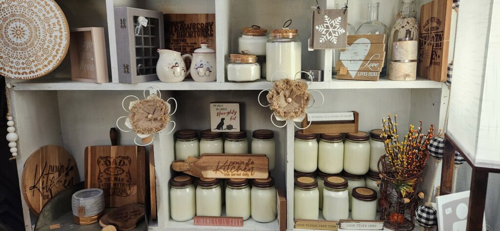 A white wooden shelf displays various white candles in glass jars, rustic wooden signs, cutting boards, ceramic jars, decorative flowers, and autumn-themed faux berry stems in a cozy, country-style arrangement.