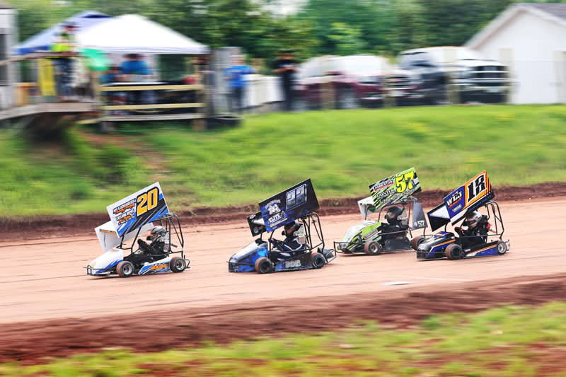 Four go-karts with colorful designs race on a dirt track at the Lake Superior Ice Festival, kicking up dust as they speed around a curve. Blurred spectators and vehicles are visible in the background.