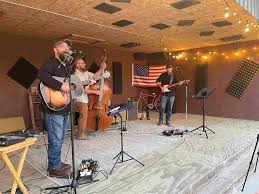 Three musicians perform on a wooden stage with guitars and a double bass. An American flag hangs behind them, and string lights decorate the ceiling. Microphones and music stands are in front of the performers.