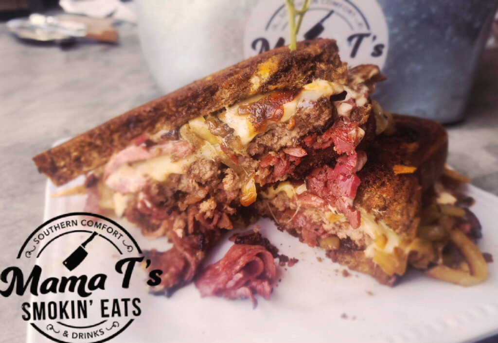 A close-up of a grilled sandwich filled with corned beef, melted cheese, and caramelized onions, cut in half on a white plate. The logo “Mama T’s Smokin’ Eats & Drinks” is featured in the corner.
