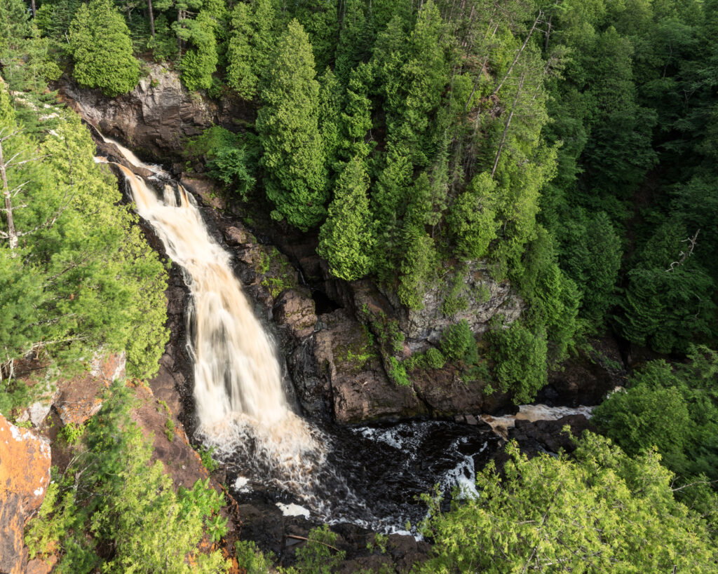 A waterfall cascades down rocky cliffs surrounded by dense forest, with white water flowing into a dark pool below—this lush landscape lies not far from famed Wisconsin snowmobile trails, blending adventure with breathtaking natural beauty.