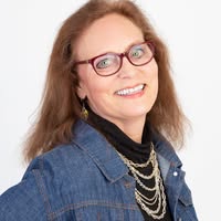 Smiling woman with glasses and long brown hair, wearing a denim jacket, layered chain necklaces, and a black turtleneck, posing against a plain white background.