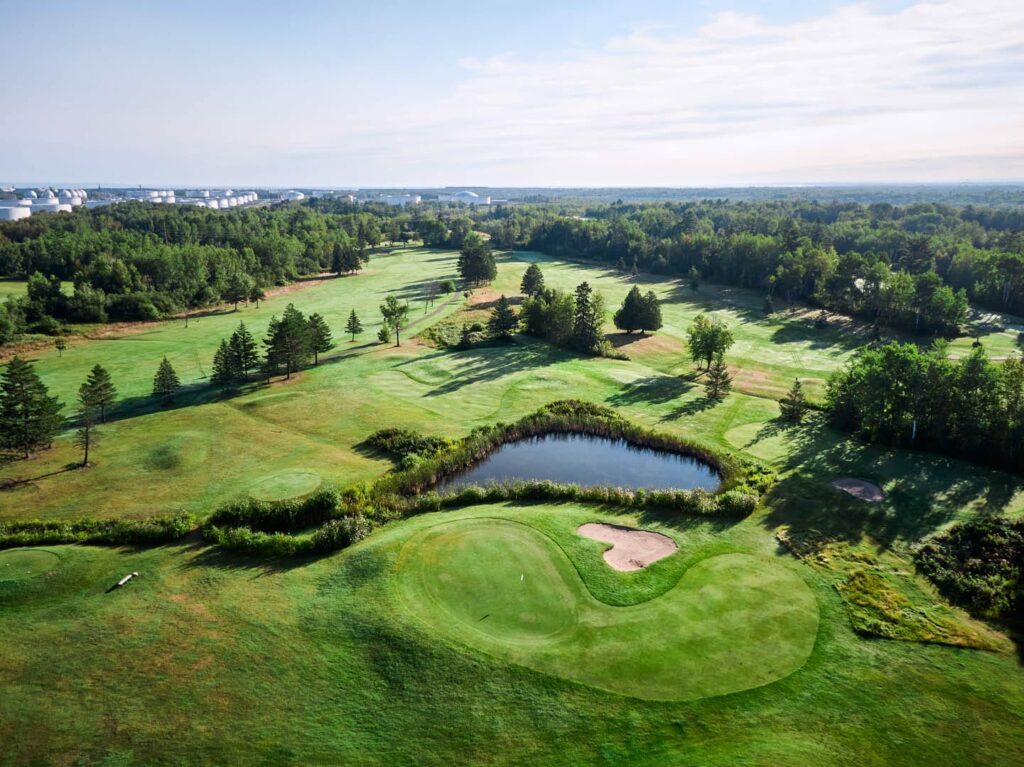Aerial view of a lush, green golf course featuring a small pond, sand bunker, and surrounding trees under a partly cloudy sky—an idyllic scene reminiscent of the scenic beauty celebrated at the Lake Superior Ice Festival.