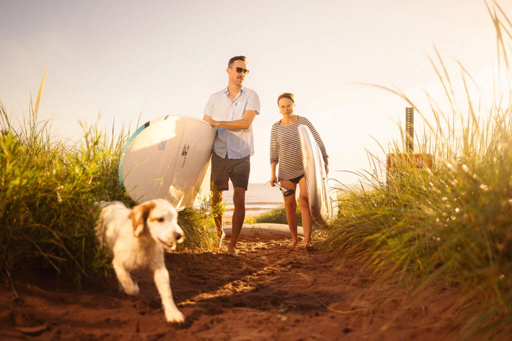 A man and woman walk along a sandy path through tall grass, each carrying a surfboard, while a white dog runs ahead toward the beach—capturing the adventurous spirit of Wisconsin places to visit in summer.