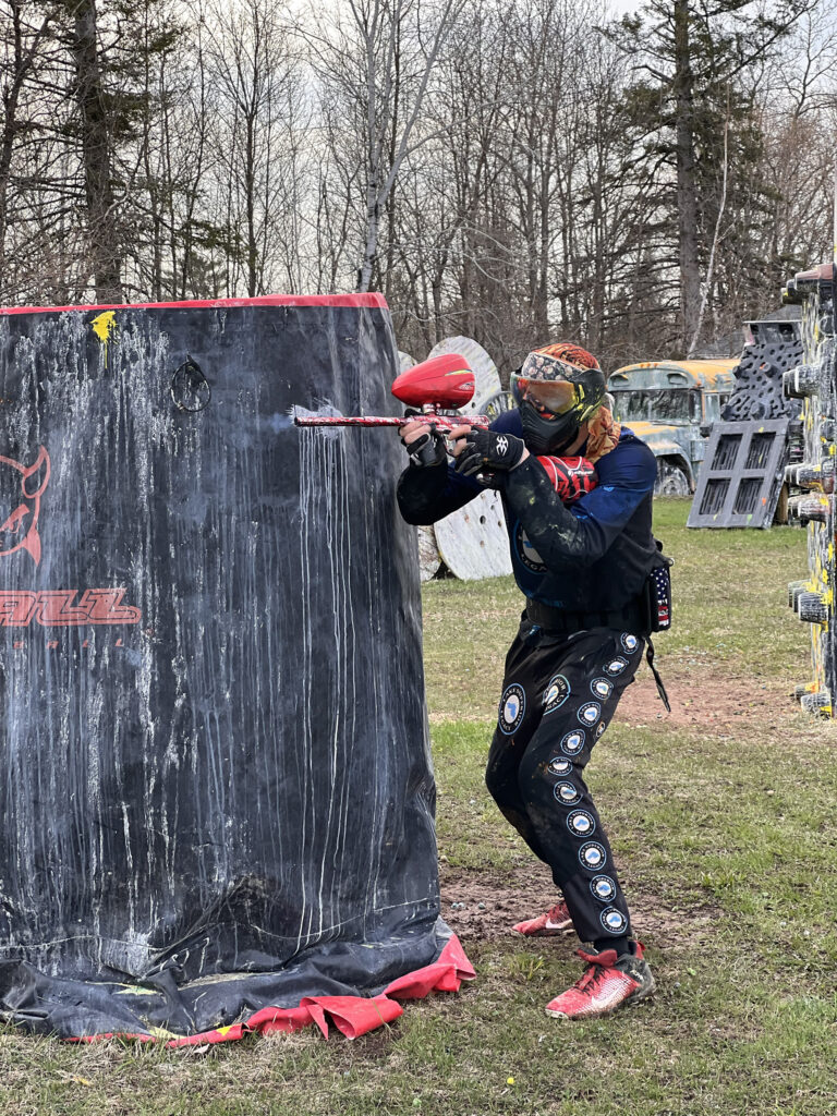 A person in paintball gear crouches beside a large black inflatable bunker, aiming a paintball marker. The ground is grassy, and bare trees are visible in the background.
