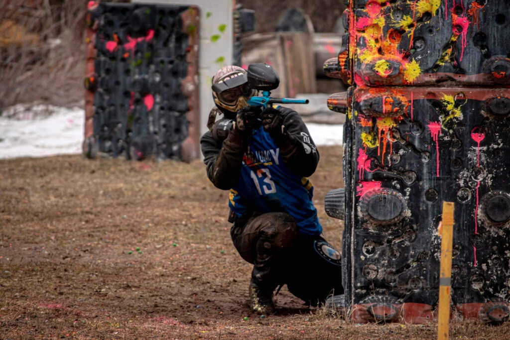 A person wearing protective gear crouches behind a paint-splattered barrier while aiming a paintball gun during an outdoor paintball game. The ground is muddy, and more barriers are visible in the background.