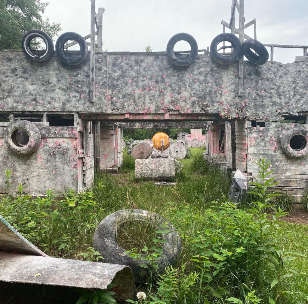 Outdoor paintball arena with weathered wooden structures, scattered tires, and splashes of pink paint. Tall grass and plants grow around the obstacles under an overcast sky.