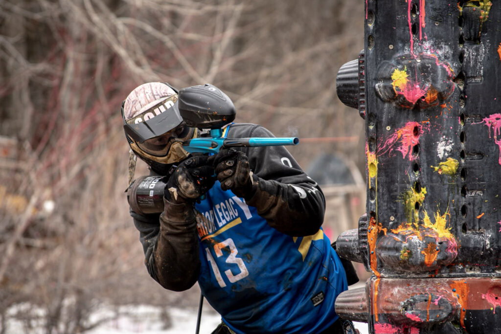 A paintball player in protective gear and a blue jersey aims a marker while taking cover behind a paint-splattered barrier during an outdoor game.