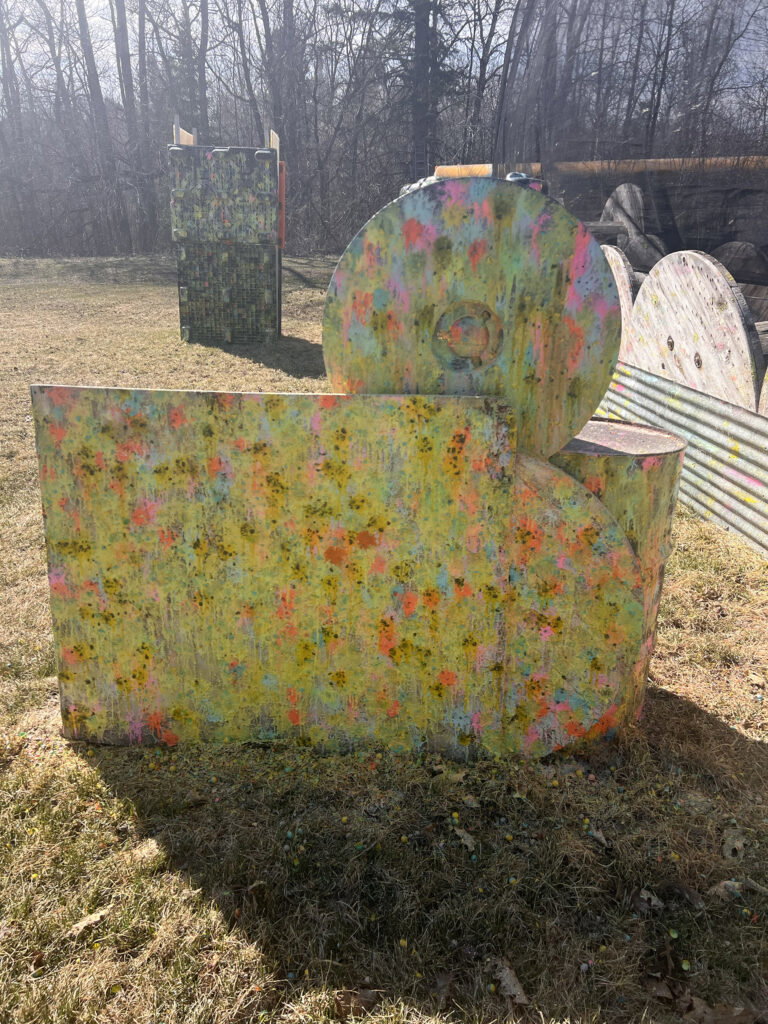Colorful, paint-splattered wooden barriers shaped like geometric forms stand on a grassy field at an outdoor paintball arena, reminiscent of the vibrant installations seen at events like the Lake Superior Ice Festival, with trees and sunlight in the background.