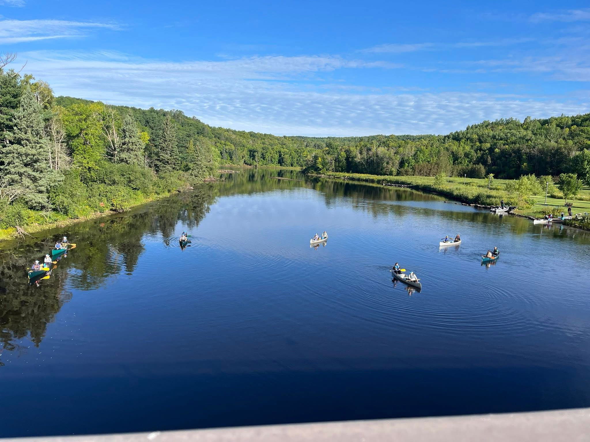 A calm river surrounded by green trees under a blue sky, with several groups of people paddling canoes on the water. The scene is peaceful and reflects nature and outdoor recreation.