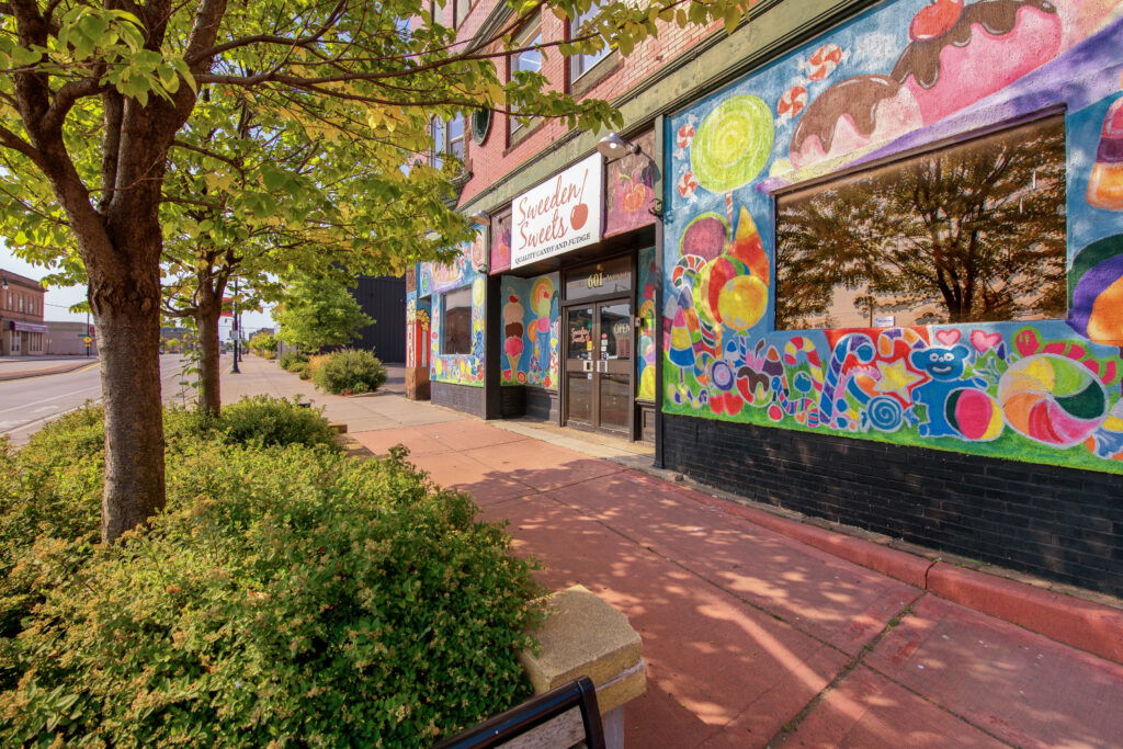 A colorful candy shop with bright murals of sweets on its windows sits along a sunny, tree-lined sidewalk in a small town, making it one of the charming Wisconsin places to visit in summer. Shrubs and a tree provide greenery in front of the store.