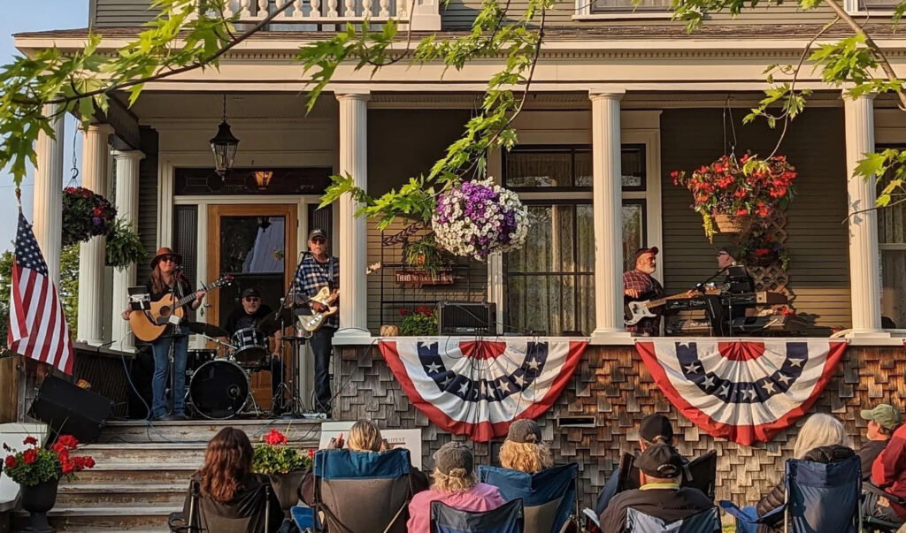 A band performs on a house porch decorated with patriotic banners and hanging flower baskets, as a seated audience enjoys the outdoor show under leafy branches—capturing the festive spirit of events like the Lake Superior Ice Festival.