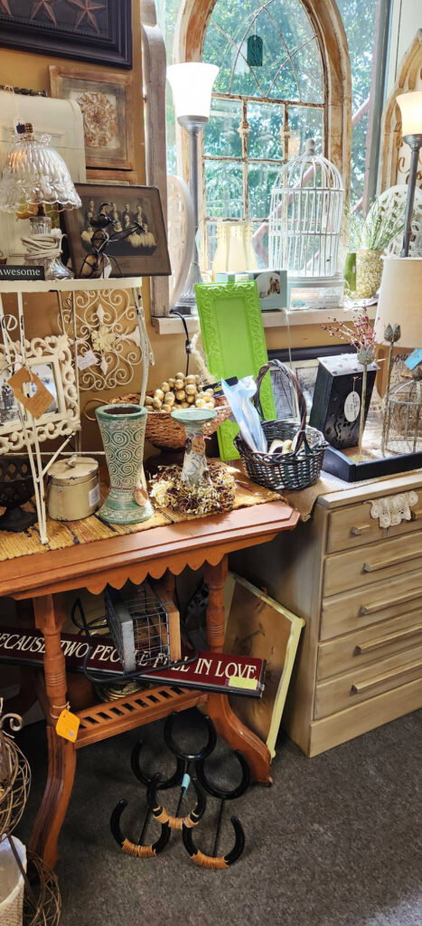 A cozy vintage shop corner displays assorted decor items, including picture frames, a clock, a lamp, vases, a woven basket, and a wooden table, with sunlight streaming through an arched window in the background.