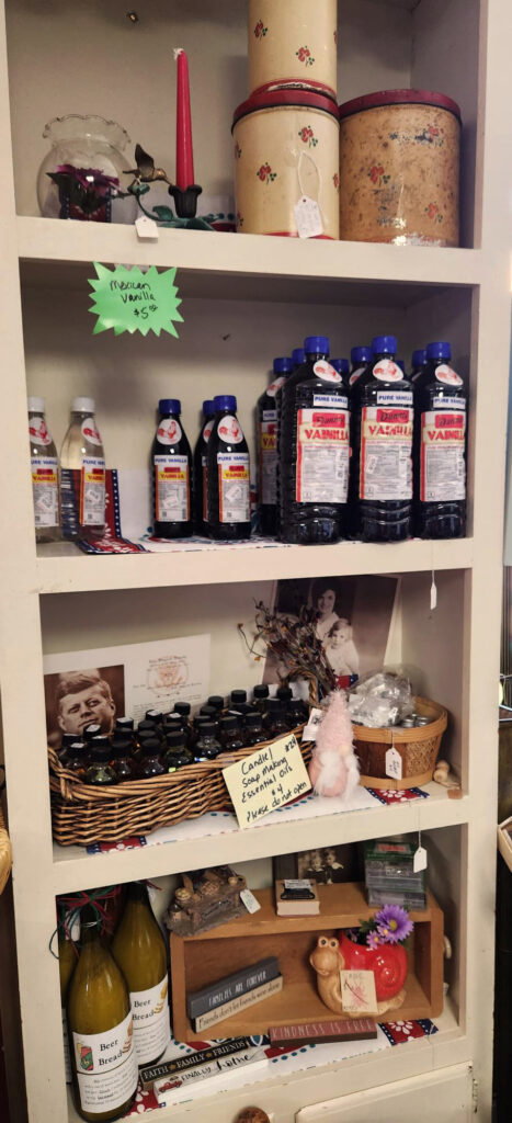A white shelving unit displays bottles of vanilla extract, jars, vintage tins, a red candle, decorative baskets, framed photos, and small trinkets, with handwritten signs indicating prices and product types.