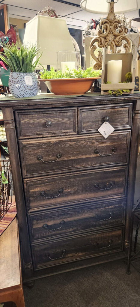 A rustic dark wood dresser with five drawers is displayed in a store. It is topped with a potted plant, a decorative candle holder, and other decor items. A price tag hangs from one of the upper drawers.