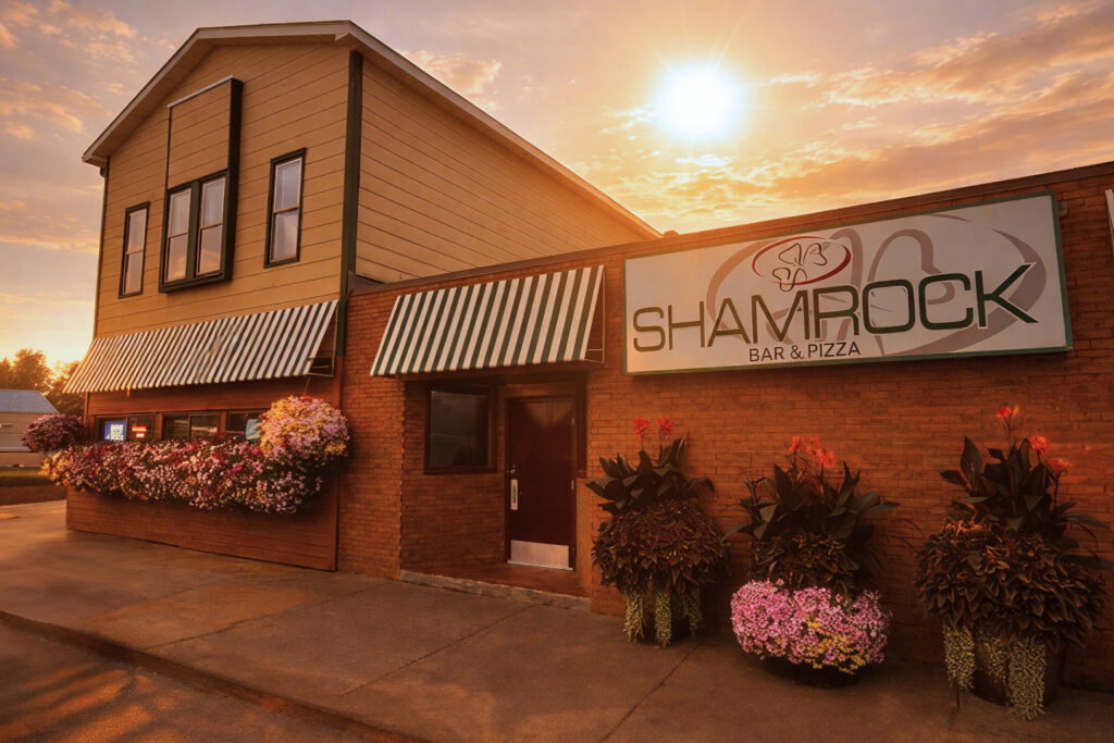 The exterior of Shamrock Bar & Pizza at sunset, with green-and-white striped awnings, flower arrangements along the building, and a large sign with the restaurant’s name on a brick wall—perfect for gathering after the Lake Superior Ice Festival.