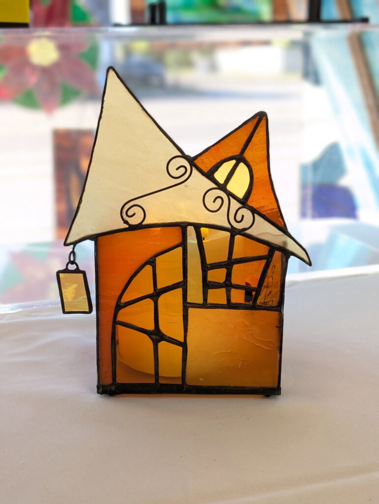 A colorful glass sculpture with abstract, geometric shapes sits on a white surface. The sculpture has orange and blue panels and black swirling line details, with a blurred background of soft greens and other colors.