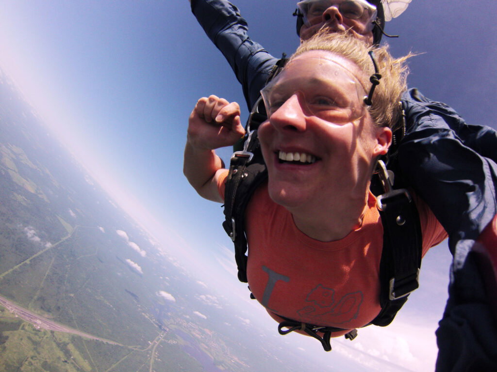 A person smiles widely while tandem skydiving, wearing a harness and orange shirt, with an instructor behind. The landscape below shows greenery, roads, and patches of clouds under a clear blue sky.