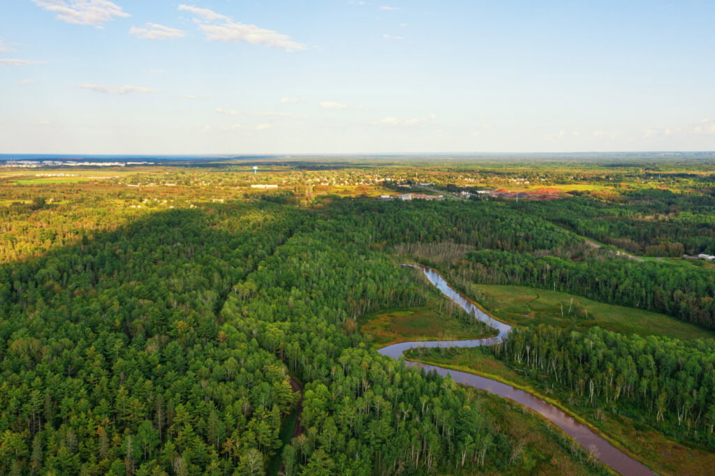 Aerial view of a lush green forest with a winding brown river cutting through the trees under a clear blue sky, with distant buildings and fields on the horizon—an inviting scene reminiscent of the Lake Superior Ice Festival's scenic beauty.