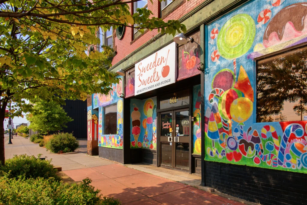 A colorful candy shop called “Sweeden Sweets” with bright murals of candies and lollipops on its exterior, located on a sunny street lined with trees and plants—one of the sweetest Wisconsin places to visit in summer.
