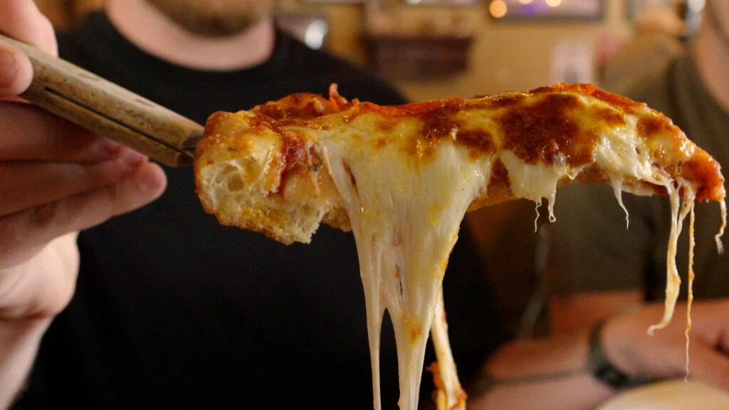 A person lifts a slice of cheesy pizza from a pie, with melted cheese stretching between the slice and the rest of the pizza—reminiscent of enjoying local eats at Wisconsin places to visit in summer. The background shows people at a table, slightly out of focus.