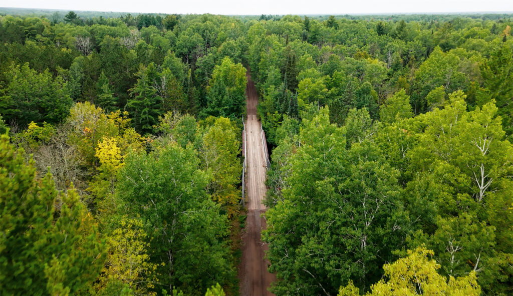 Aerial view of a narrow dirt road cutting through a dense, green forest with tall trees on both sides, resembling Wisconsin snowmobile trails, and a wooden bridge spanning a small gap in the middle of the scene.