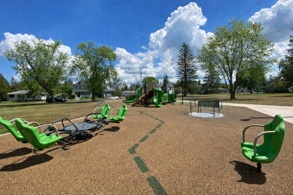 A playground with green equipment, including a merry-go-round, accessible spinner, and slide structure, is set in a park with trees and grass near houses—perfect for family fun during the Lake Superior Ice Festival under a blue sky with fluffy clouds.