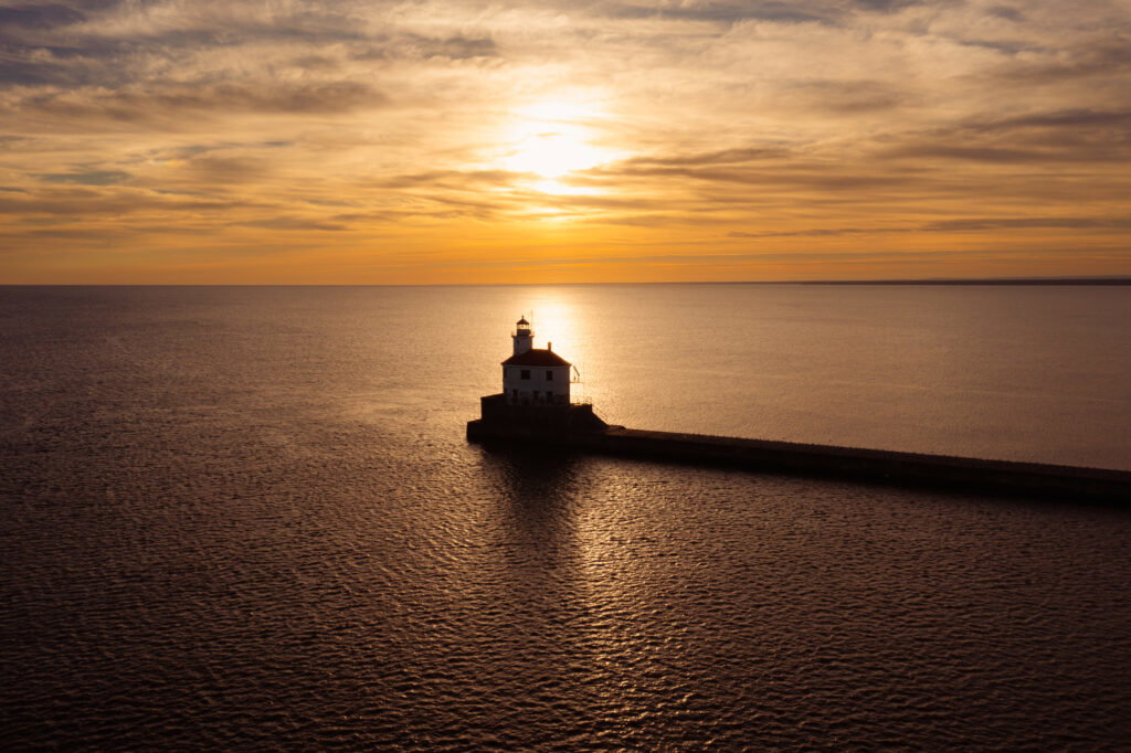 A lighthouse stands at the end of a long pier, surrounded by calm water, as a golden Lake Superior Ice Festival sunset casts warm light and shimmering reflections across the sky and sea.