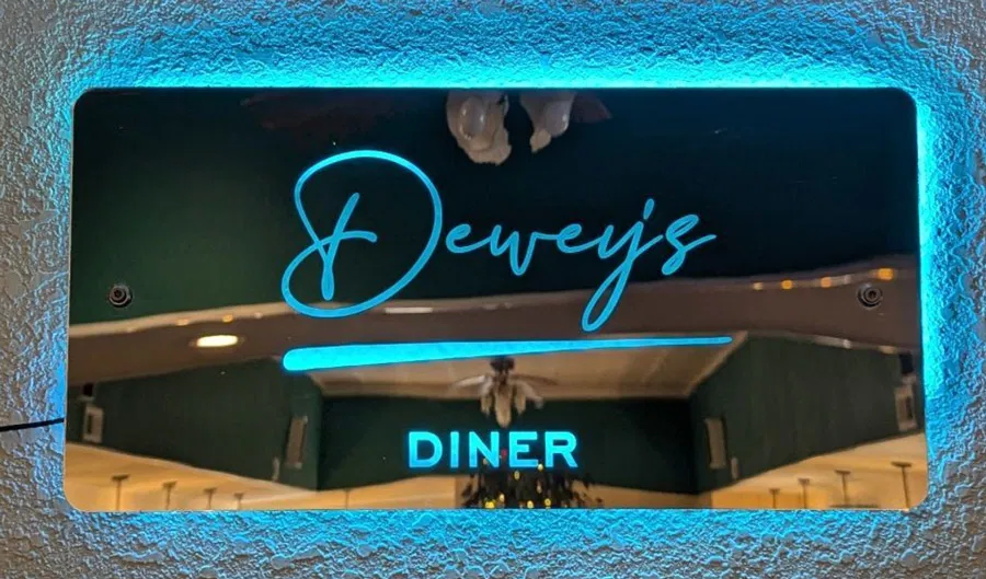 A glowing neon sign reads "Dewey's DINER" in blue cursive and block letters, mounted on a textured wall with ceiling lights and a fan reflected below the sign.