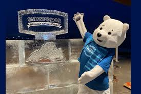 A person in a polar bear costume wearing a blue shirt poses with an ice sculpture that reads "SNOWFEST Manitowish Waters Chamber." The background is dark, suggesting the event is taking place at night.