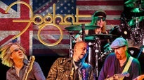 Rock band Foghat performing on stage; three musicians play guitar in front while a drummer is in the back. The British and American flags are displayed behind them, with the Foghat logo overlaid.