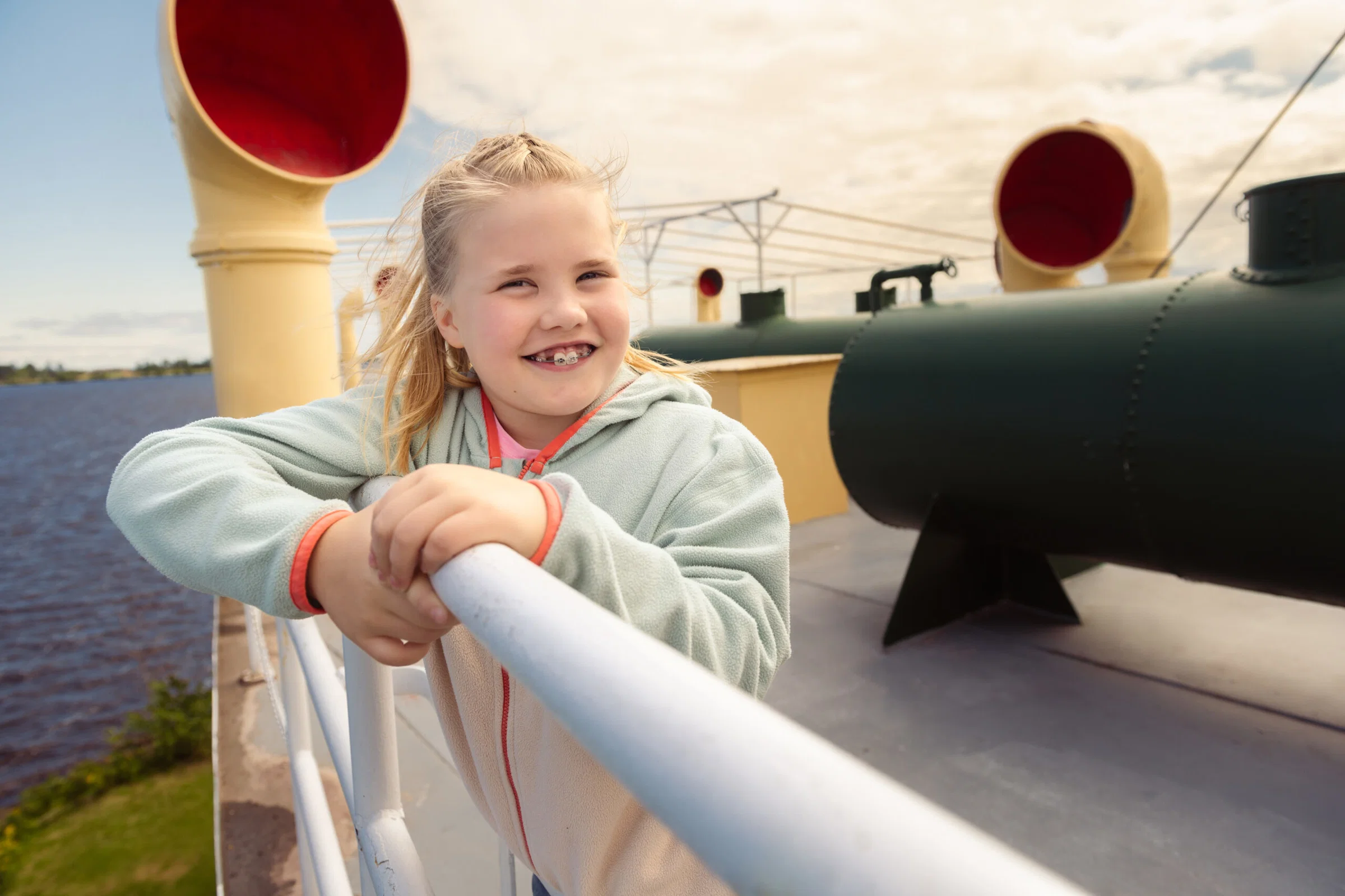 25% center A smiling girl with blonde hair and braces leans on a metal railing outdoors, with large ship funnels and a river—one of the memorable Wisconsin places to visit in summer—visible in the background under a partly cloudy sky.