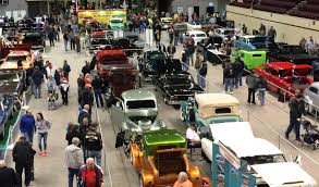 A busy indoor car show with rows of classic and vintage cars on display. Many people are walking around, viewing the vehicles inside a large well-lit exhibition hall.