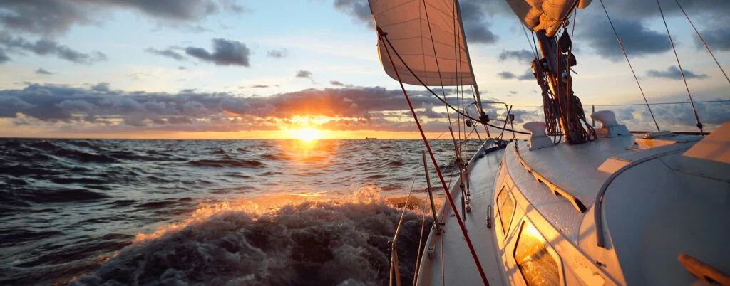 A sailboat glides through ocean waves at sunset, with the sun low on the horizon, casting warm light on the boat and clouds—a perfect scene for those seeking outdoor activities in Superior Wisconsin.